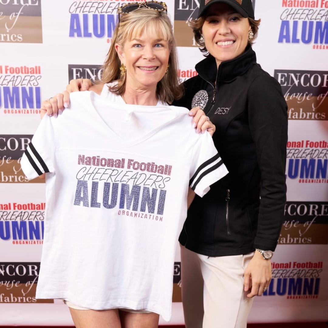 Two women holding a white shirt with 'National Football Cheerleaders Alumni' text against a branded backdrop.